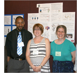 Three adults standing next to one another, in front of a presentation board, smiling at the camera.