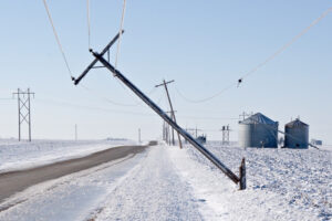 A power pole weighed down by ice accumulation bends over a snow-covered road.